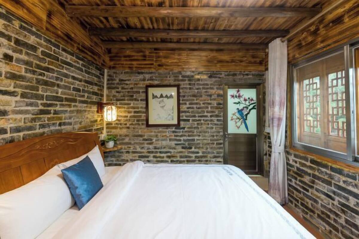 A room with a stone wall, wooden ceiling, a bed with white bedding, a framed picture on the wall, and a window with curtains.
