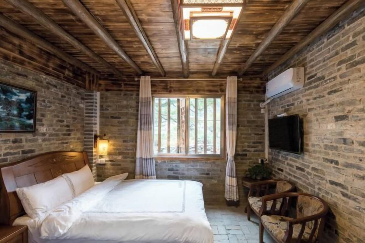 A room with a wooden ceiling, a bed with white linens, a TV mounted on the wall, and a window with curtains.