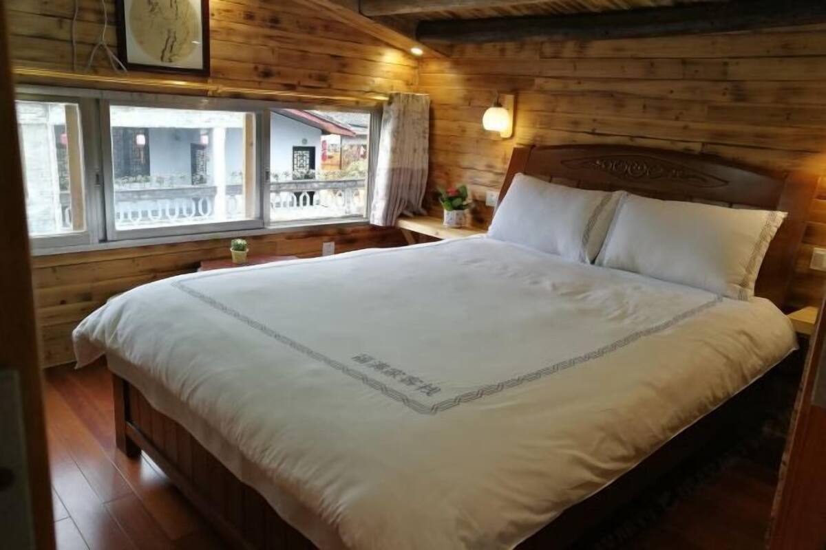A wooden-paneled room with a large bed, a window with a view, and a small table with a plant.