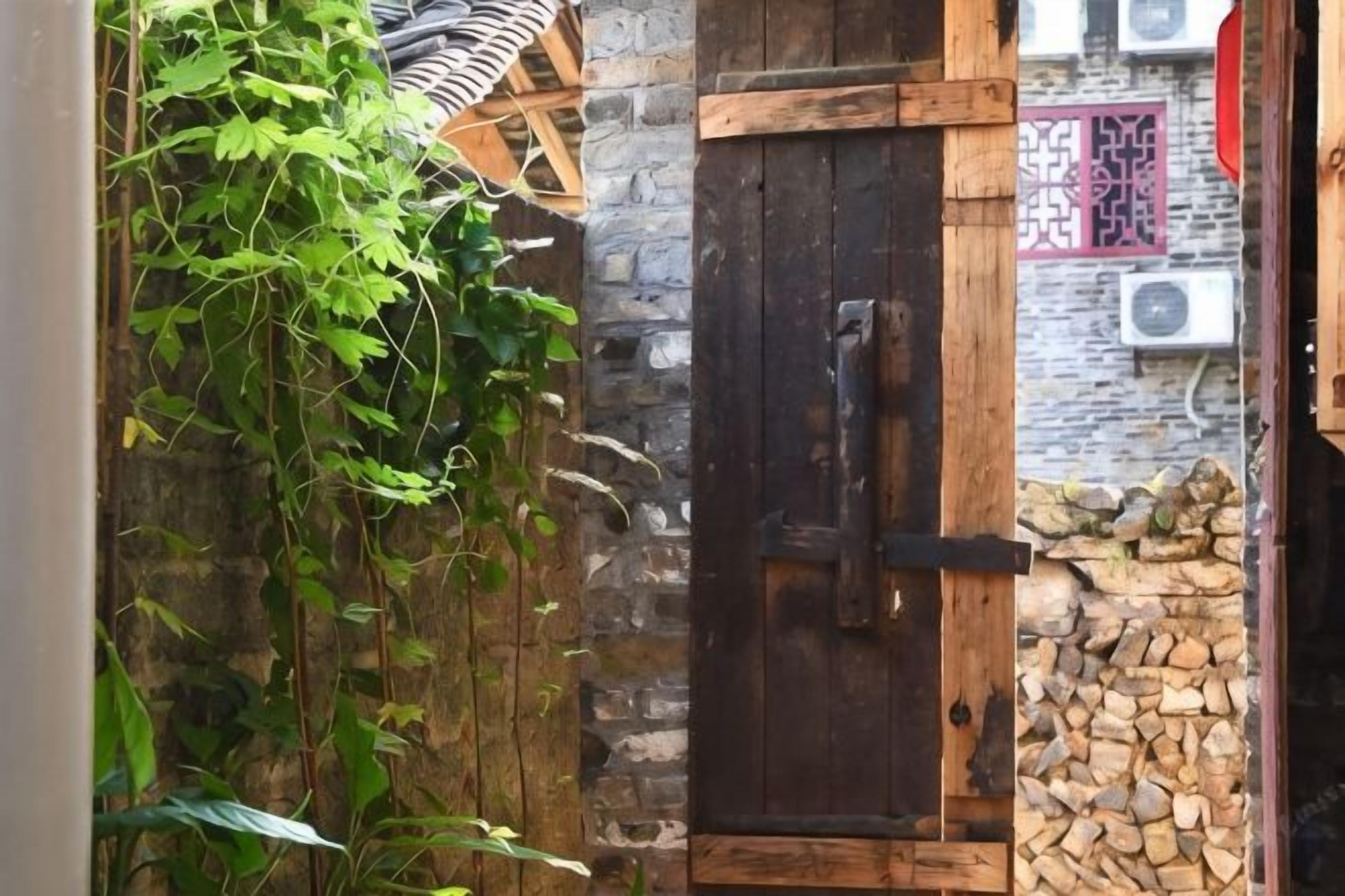 Uma porta de madeira rústica com uma tranca de metal, parede de pedra e uma pilha de lenha à direita.