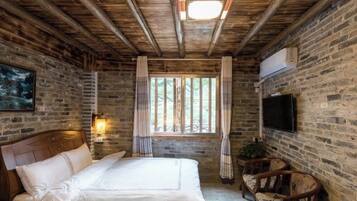 A bedroom with a wooden ceiling, brick walls, a bed with white linens, a TV mounted on the wall, and a window with curtains.