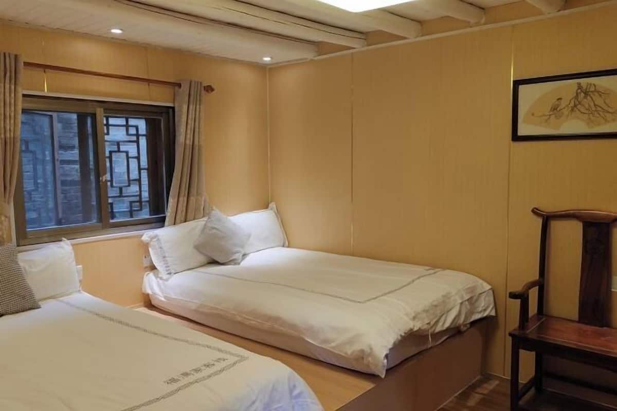 A room with two beds, a wooden chair, a window with curtains, and a framed picture on the wall.