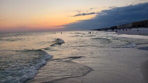 Una playa con olas al atardecer, un paseo marítimo con edificios y gente caminando.