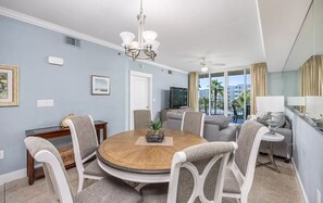 A modern living room with a round wooden table, grey upholstered chairs, a chandelier, a flat-screen TV, and a view of palm trees through large windows.