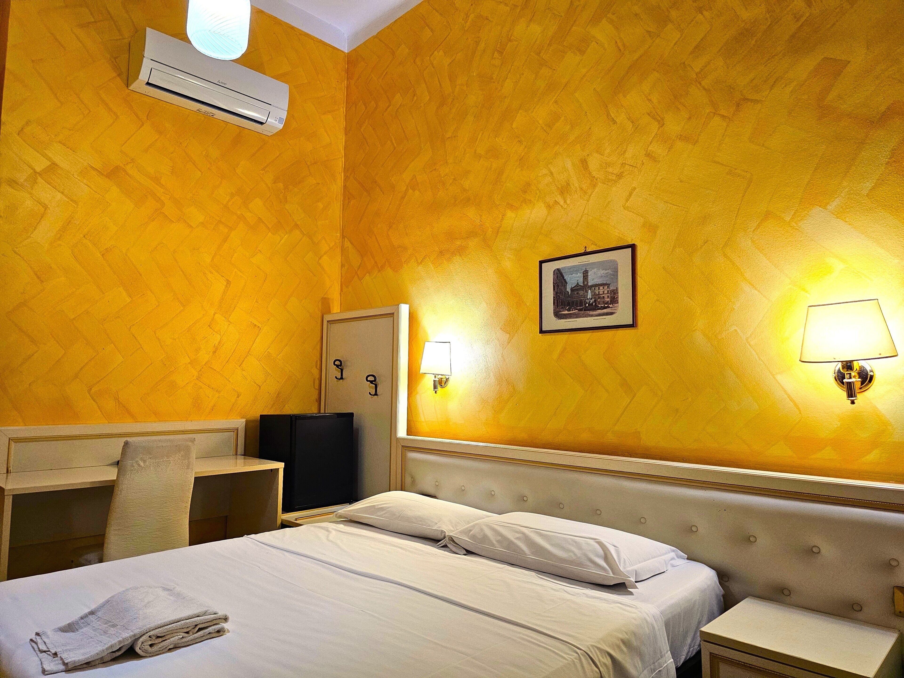 A hotel room with a bed, bedside tables, a desk, an air conditioner, and a wall-mounted lamp.
