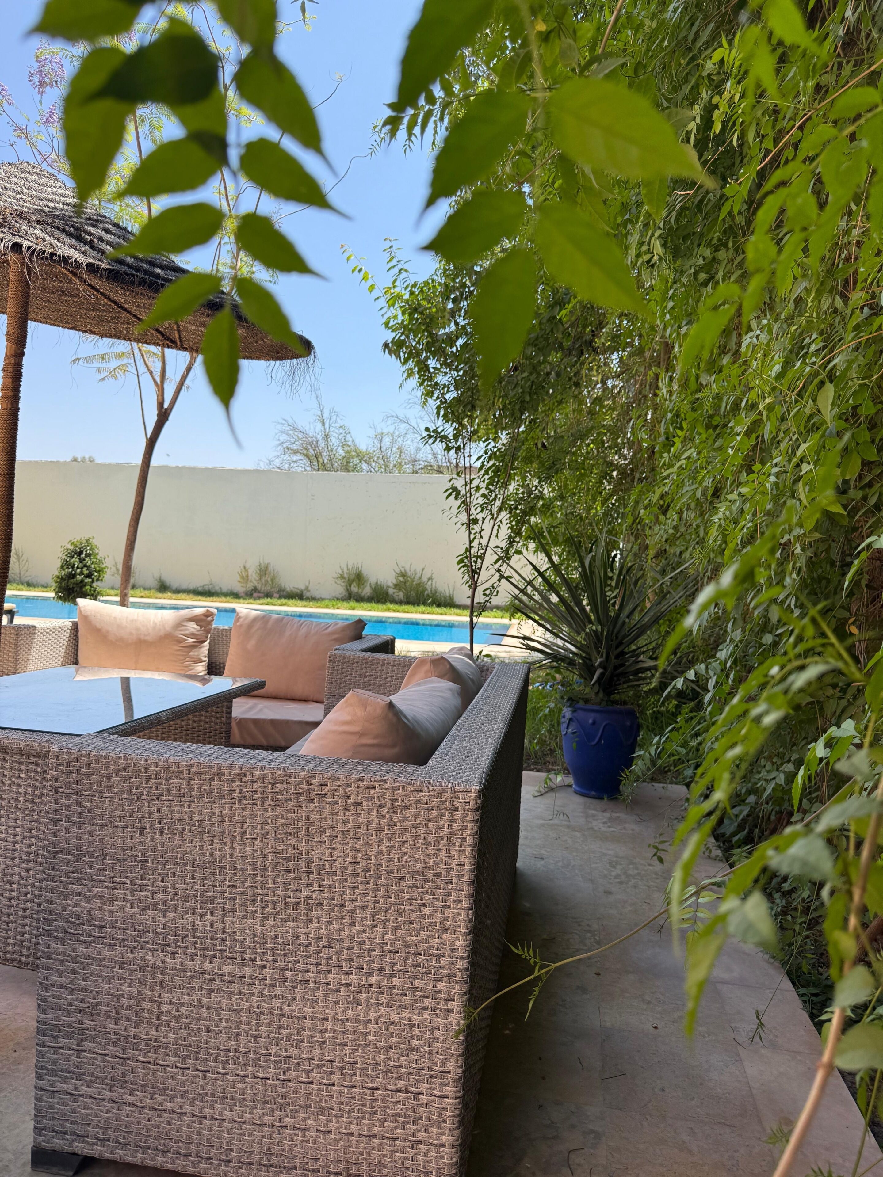 A wicker outdoor seating area with cushions, a pool, and lush greenery.
