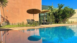 A clear swimming pool with lounge chairs and umbrellas, surrounded by greenery and a building facade.