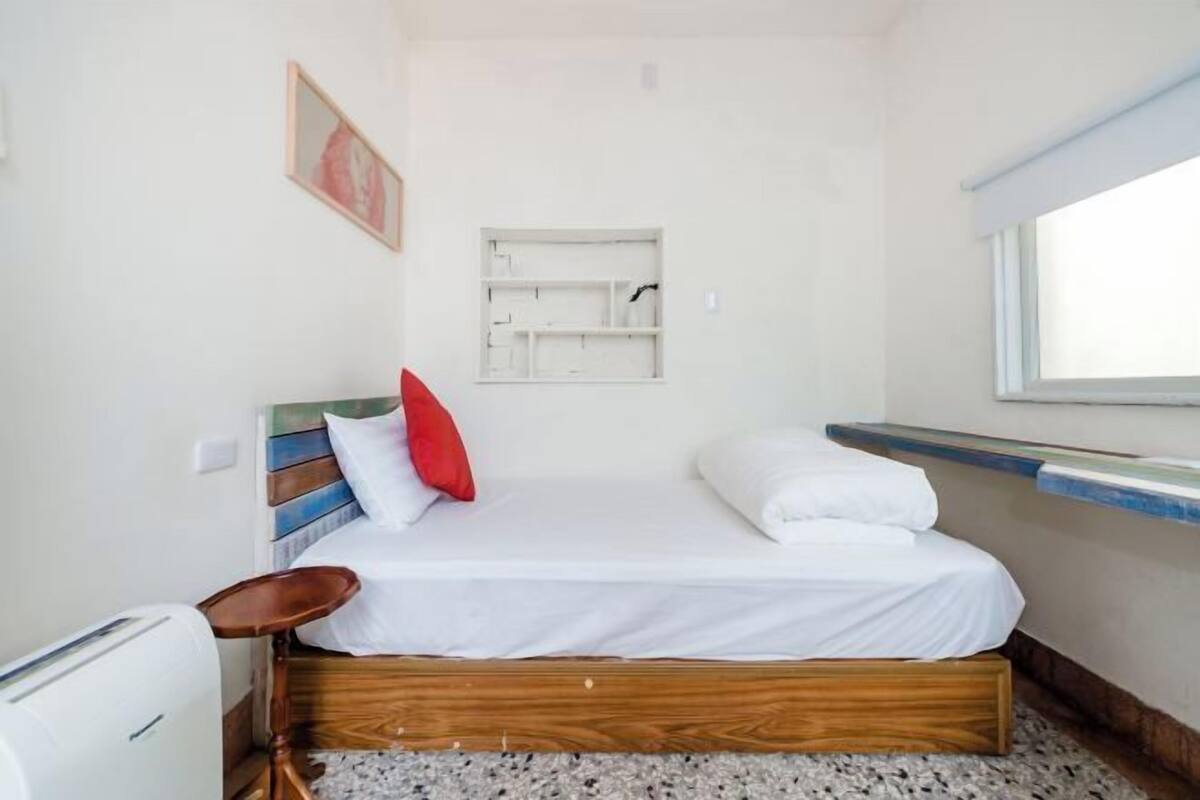 Un dormitorio con cama de madera, ropa de cama blanca, almohada roja, una pequeña silla, estante empotrado y una ventana.