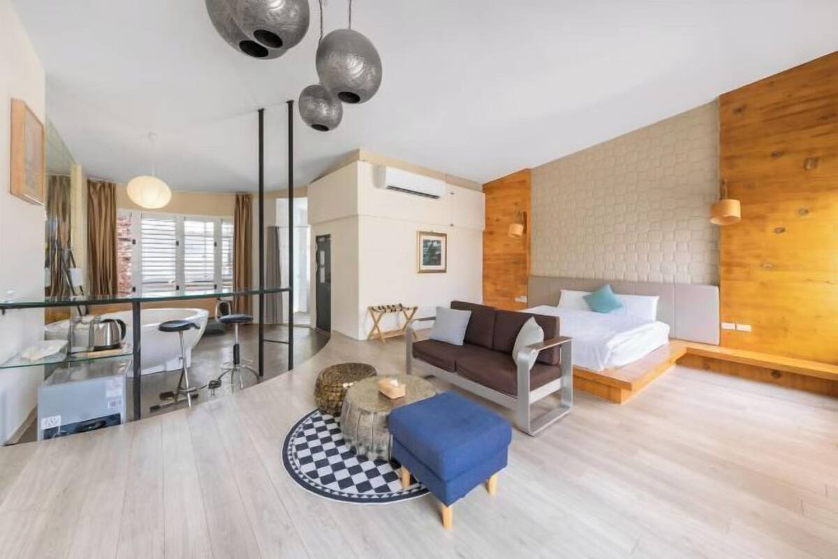 Una sala moderna con sofá, una mesa de centro y un tapete redondo. Se ve una cama con ropa de cama blanca y cabecera de madera. Un espejo grande cuelga de la pared y hay una ventana con persianas.