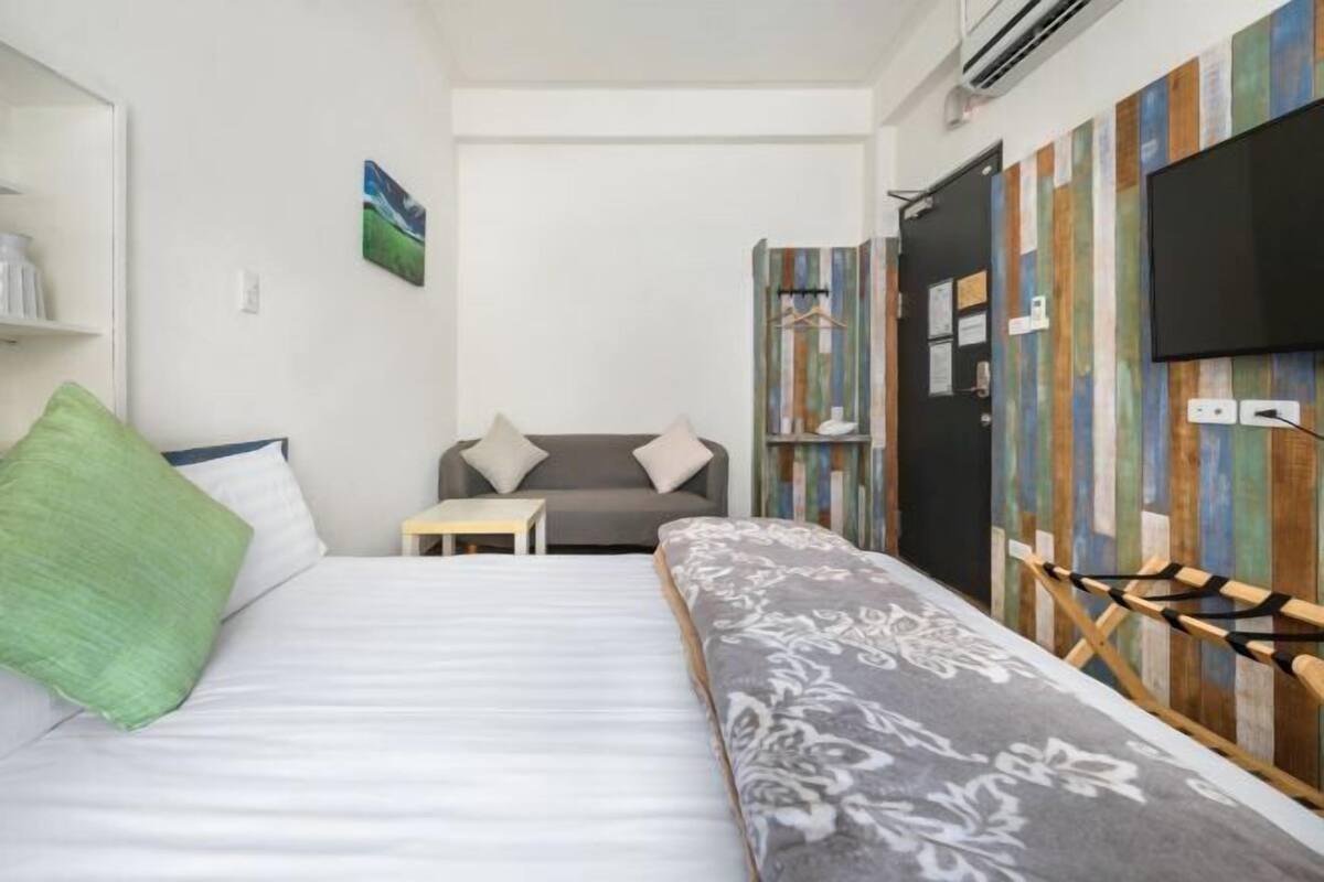 Un dormitorio con cama, una mesita, un sofá y una televisión instalada en una pared de paneles de madera.