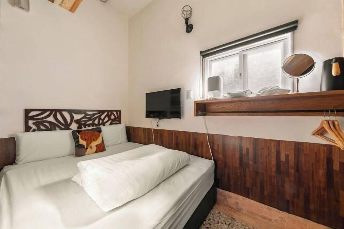 Un dormitorio con cama, televisión montada en la pared, ventana y cabecera de madera.