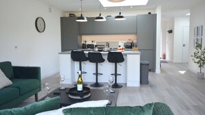 Een moderne woonkamer met een groene bank, een bijzettafeltje met wijnglazen, en een keuken met grijze kastjes en een centraal eiland.