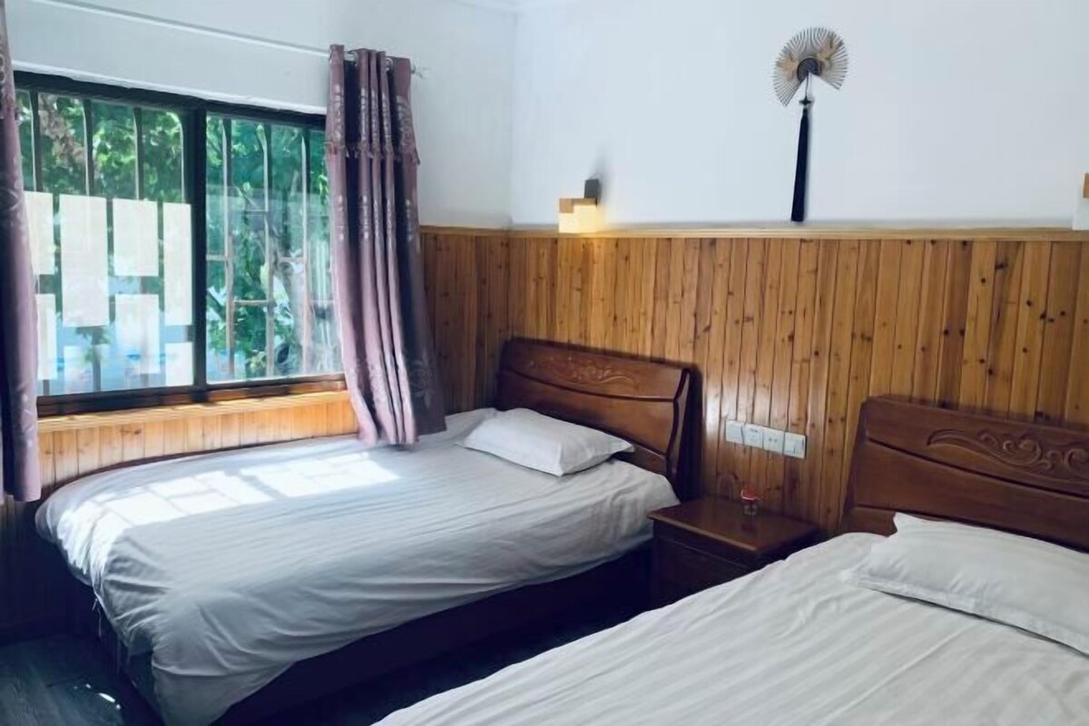 A room with two beds, a wooden wall paneling, a window with curtains, a bedside table, and a wall-mounted fan.