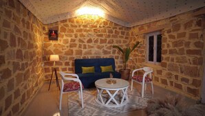 A cozy room with a stone wall, a blue sofa with yellow cushions, two white chairs, a small round table, and a potted plant. - Teela Jaisalmer Luxury Desert Swiss Tent (Jaisalmer)
