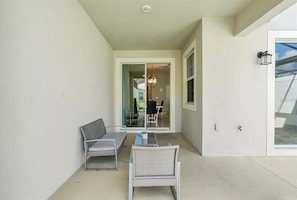 A balcony with a table and two chairs, a sliding glass door leading to an indoor area, and a wall-mounted light fixture.