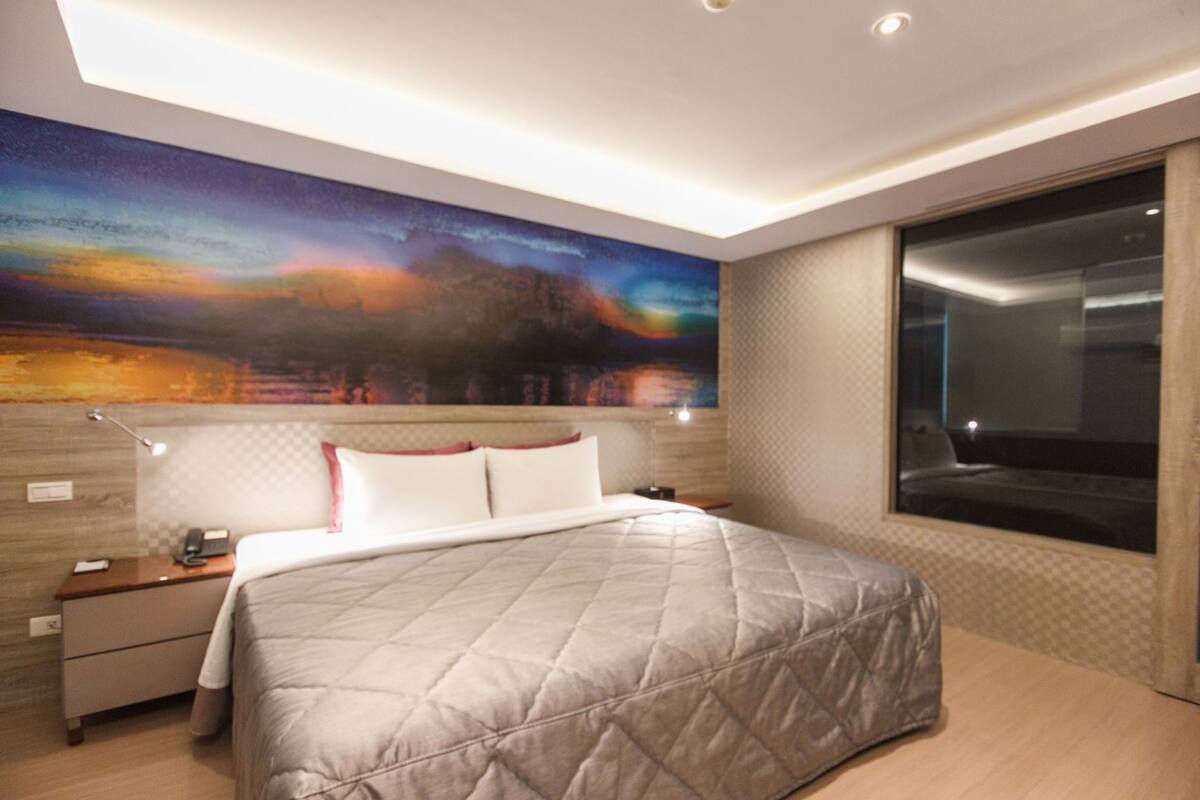 Una habitación de hotel moderna con una cama grande, una mesita de noche, un escritorio, un ventanal con vistas y un mural en la pared.