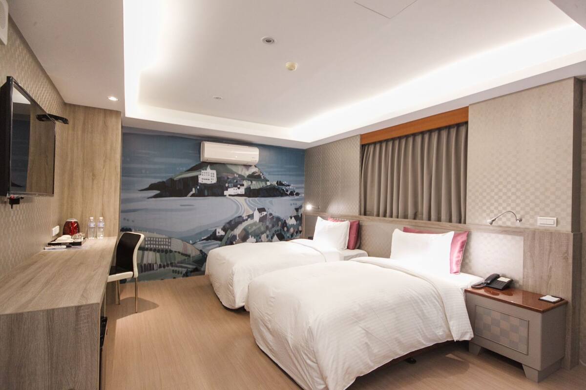 Habitación de hotel con dos camas, un escritorio, una silla, una mesita de noche y aire acondicionado instalado en la pared.