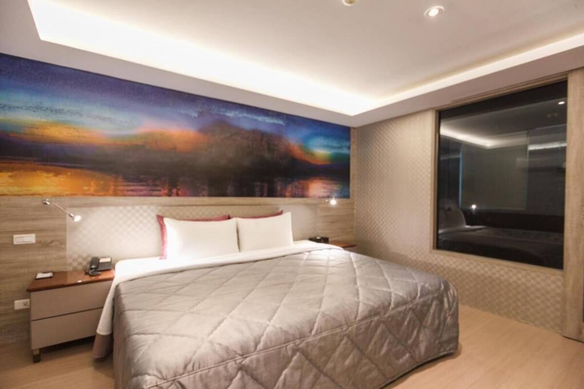 Una habitación de hotel moderna con una cama grande, una mesita de noche, un escritorio y un ventanal amplio.