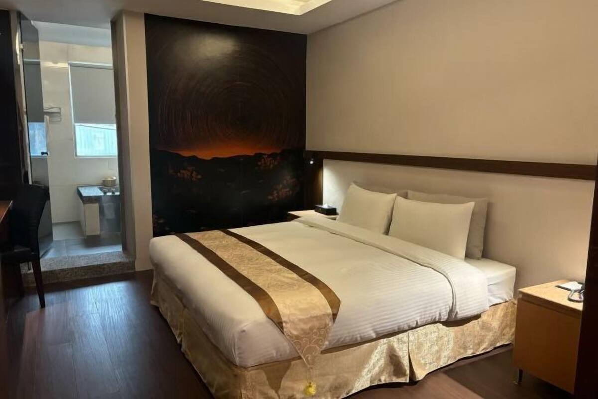 Habitación de hotel con una cama grande, cabecera, mesita de noche, escritorio y vista a un paisaje urbano a través de la ventana.