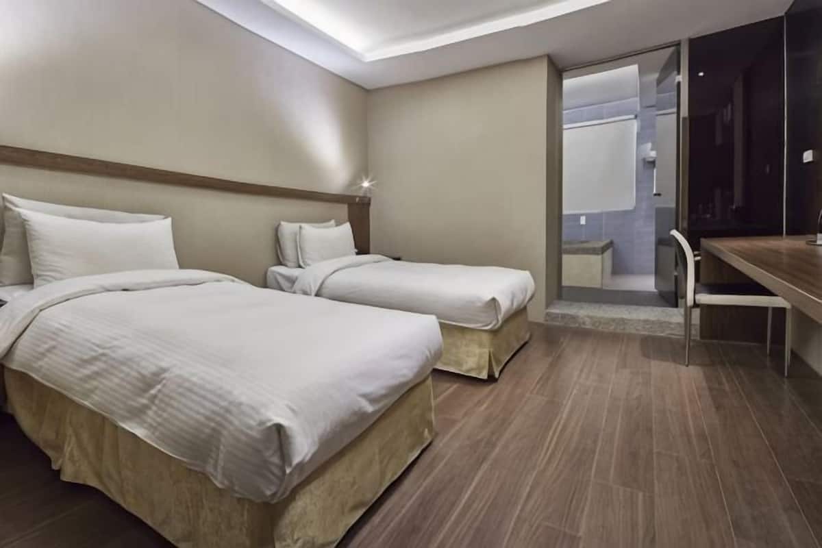 Habitación de hotel con dos camas, un escritorio, una silla y zona de ducha.