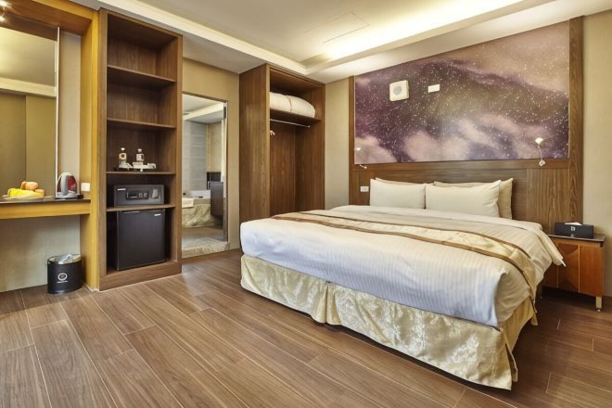 A hotel room with a large bed, wooden wardrobe, a small desk, and a wall-mounted TV.