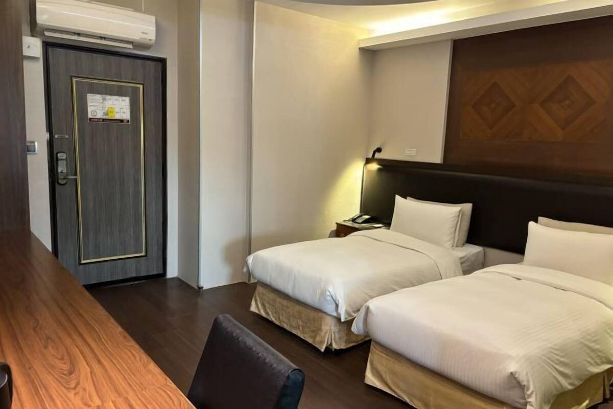 Habitación de hotel con dos camas, un escritorio de madera, una puerta con un aviso y un aire acondicionado instalado en la pared.
