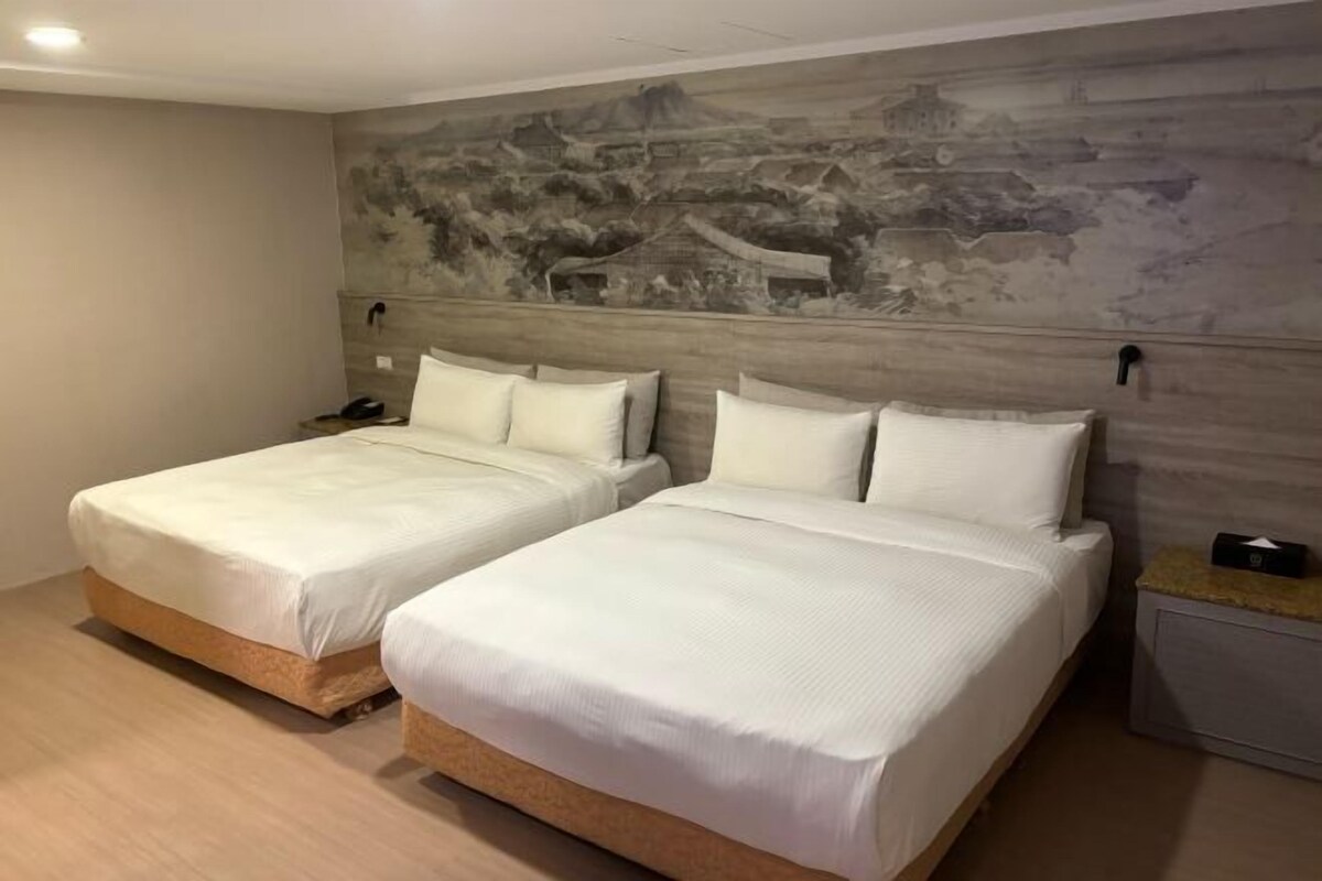 A hotel room with two beds, a wooden headboard, a wall mural, and a nightstand with a lamp.
