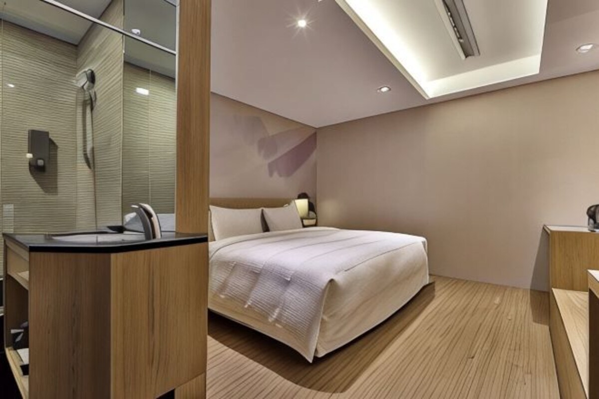 A modern hotel room with a wooden floor, a bed with a white bedspread, a bathroom with a mirror, and a nightstand.