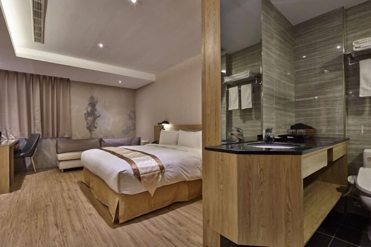 A modern hotel room with a bed, a desk, a sofa, a bathroom with a mirror, and a toilet.