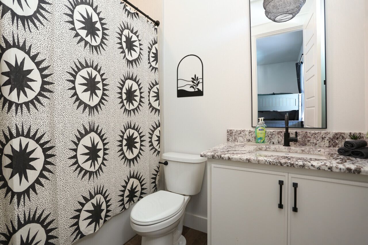 A bathroom with a sunburst shower curtain, a toilet, a sink with a mirror above it, and a small window.
