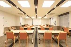 Un salón de conferencias con varias mesas y sillas, una pantalla de proyector y un amplio ventanal con persianas.