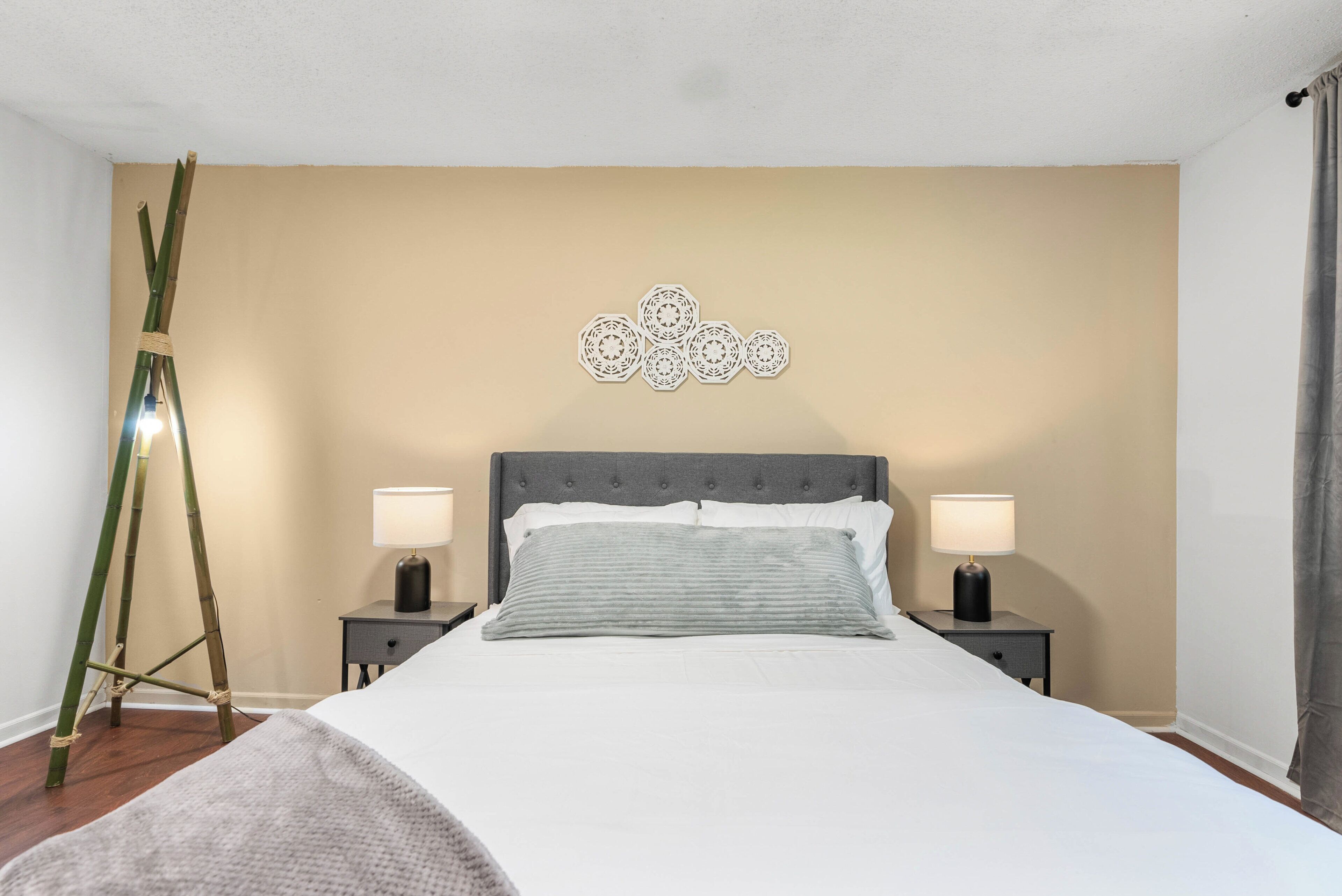 A neatly made bed with a grey headboard, two bedside tables with lamps, a wall-mounted decorative piece, and a bamboo ladder.