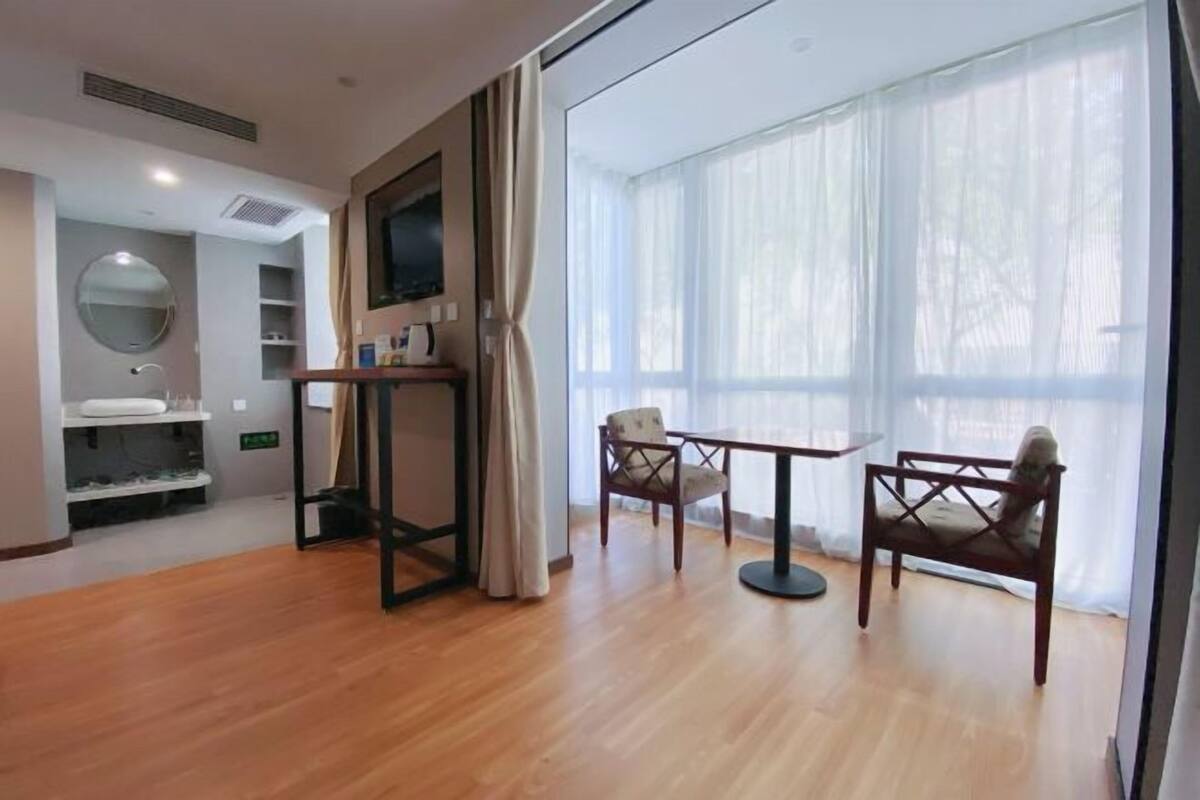 Habitación de hotel con piso de madera, un espejo redondo, televisor montado en la pared, una mesita con una silla y un amplio ventanal con cortinas traslúcidas.