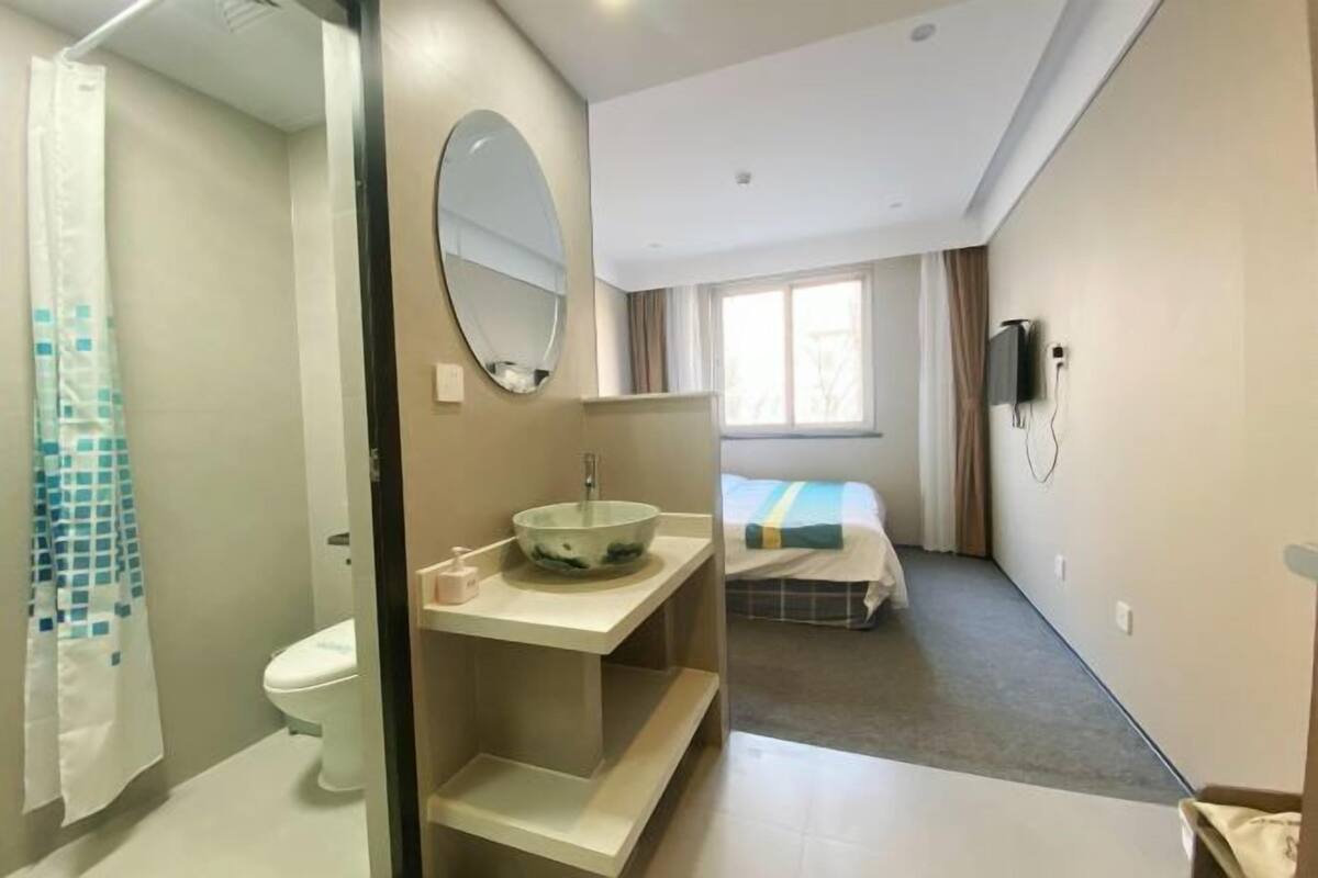 Un baño con inodoro, lavamanos y espejo. Hay una cortina de ducha, una cama con colcha azul y blanca a rayas, y una ventana con cortinas.