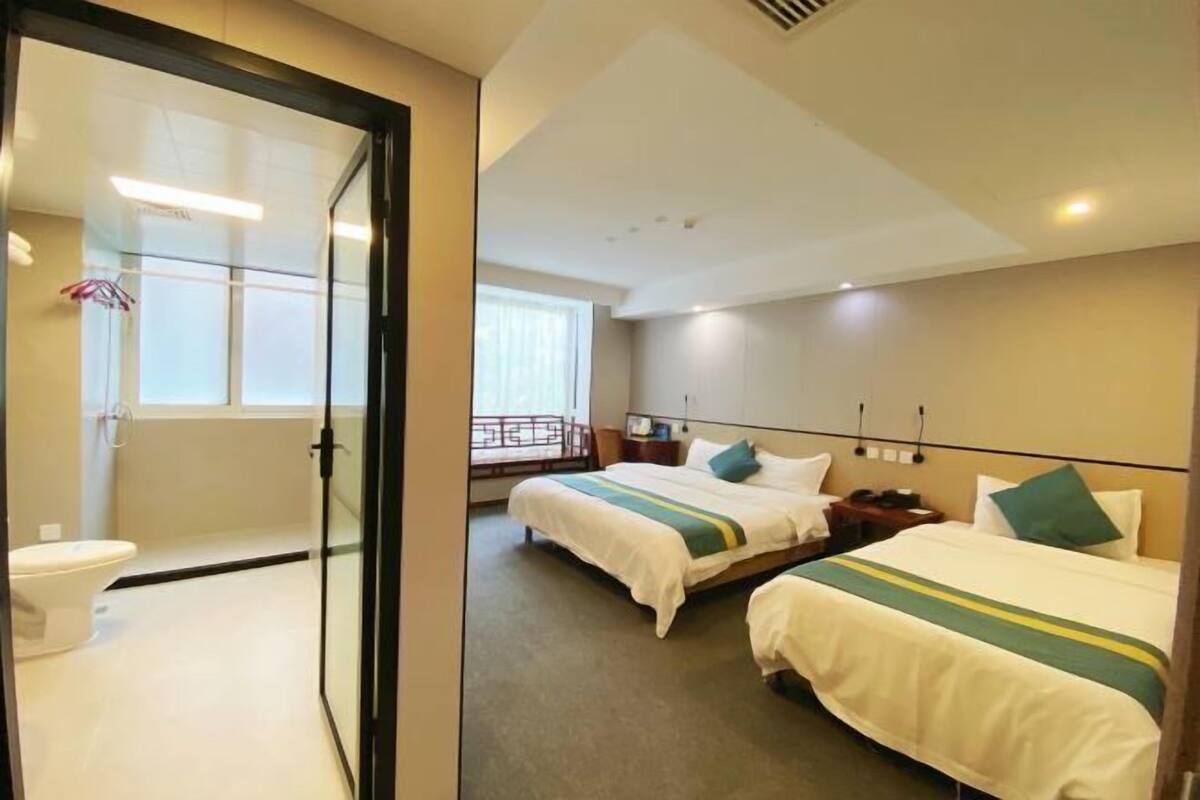 Habitación de hotel con una mampara de vidrio, una cama con cubrecama verde y blanco a rayas, una mesita de noche con lámpara y una ventana con cortinas.