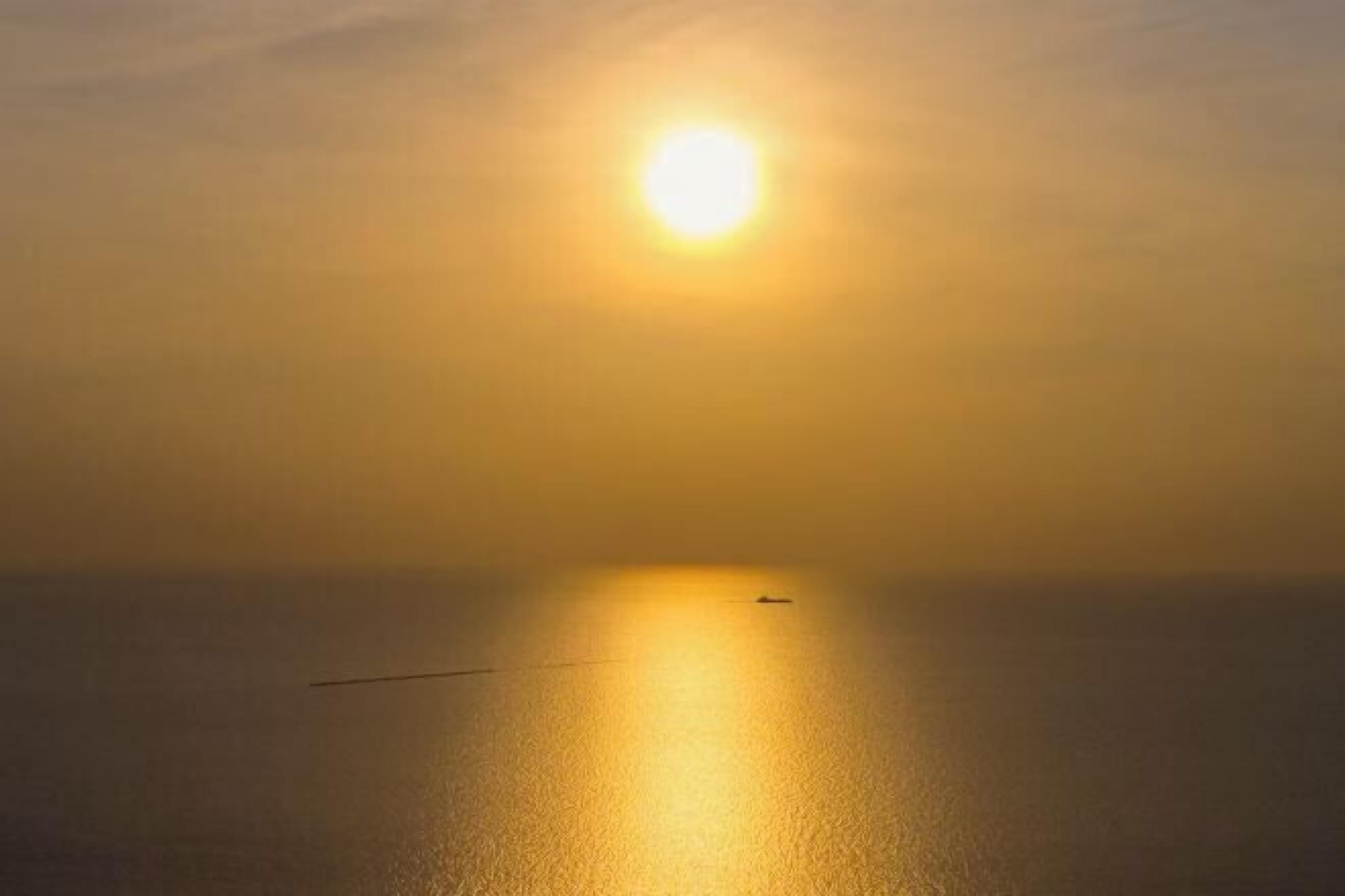A calm sea with a boat in the distance and a bright sun above.