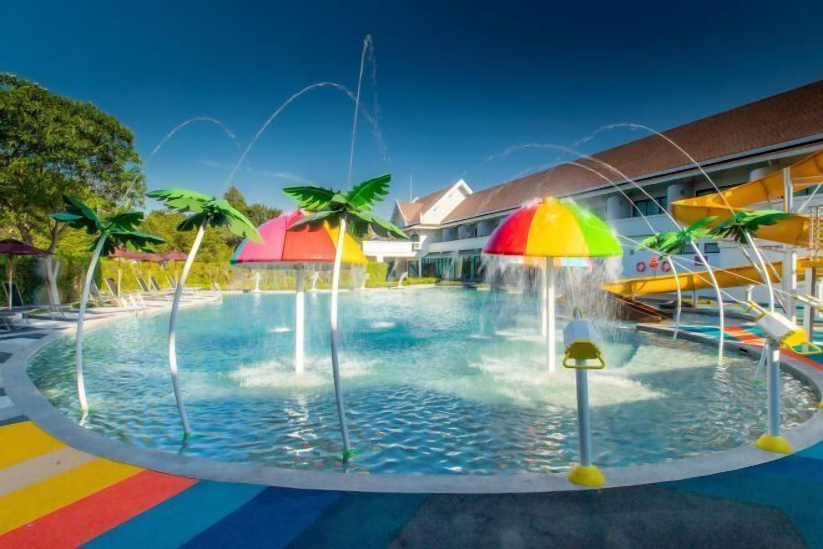 A circular pool with water jets, surrounded by colorful umbrellas and palm trees.
