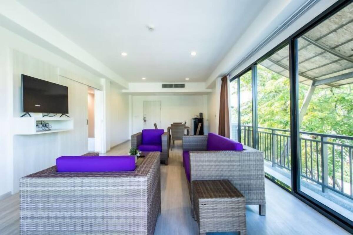 A modern living room with wicker furniture, a flat-screen TV, and a balcony with a view of greenery.