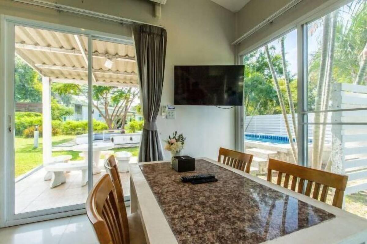 A room with a dining table, chairs, a TV mounted on the wall, and a sliding glass door leading to an outdoor area with a patio and greenery.