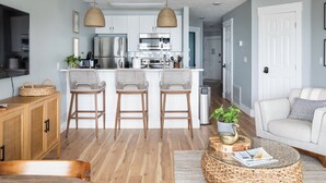 Une cuisine moderne avec des meubles blancs, des appareils en acier inoxydable et un îlot central doté de tabourets de bar. On y trouve également un coin repas avec un canapé blanc et une table basse en verre.