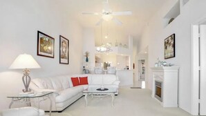 A modern living room with a white sofa, a glass coffee table, and a ceiling fan. There is a fireplace on the right and a kitchen area in the background. - " SPANISH WELLS GOLF & CC "     Chilled Pool for summer fun! (Bonita Springs)