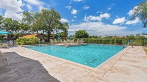 A rectangular swimming pool with a tiled deck, surrounded by lounge chairs and umbrellas, with trees and a clear sky in the background. - " SPANISH WELLS GOLF & CC "     Chilled Pool for summer fun! (Bonita Springs)