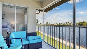 A balcony with a view of a body of water, a covered seating area with blue cushions, and a glass door leading to a bedroom. - " SPANISH WELLS GOLF & CC "     Chilled Pool for summer fun! (Bonita Springs)