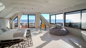 Ein modernes Wohnzimmer mit einem großen Bett, einer Couch, einem Sitzsack und einem Couchtisch. Große Fenster bieten Blick auf den Ozean und es gibt einen Balkon.