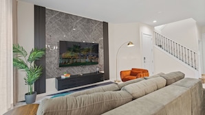 Un salon moderne avec un grand téléviseur à écran plat fixé sur un mur en marbre, un canapé d’angle, une lampe sur pied et un escalier en arrière-plan.