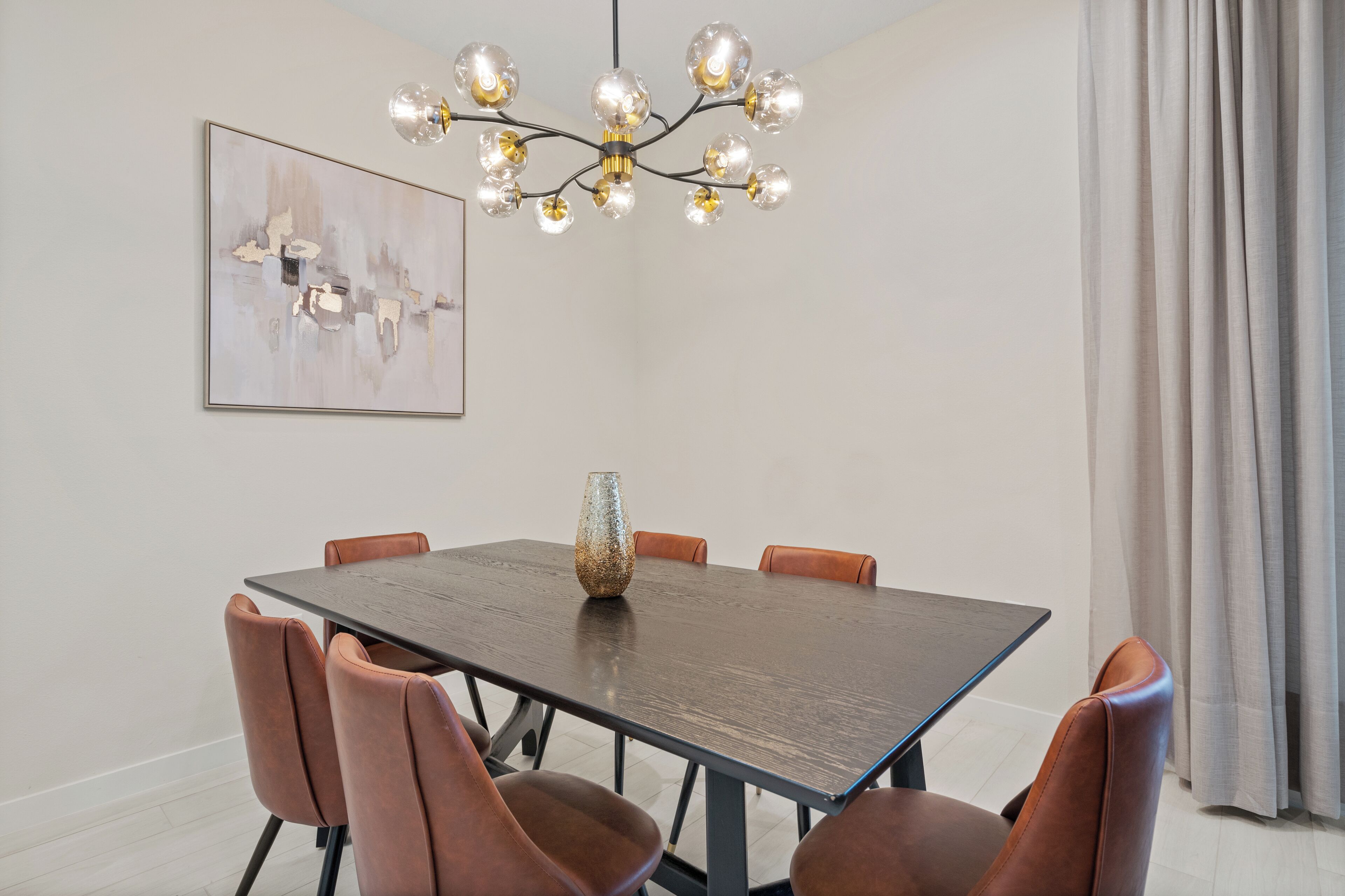 A dining area with a rectangular table, six brown chairs, a decorative vase, a chandelier, and a framed abstract painting on the wall.