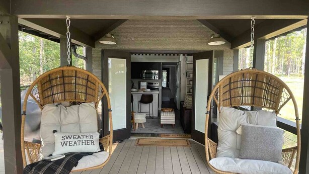 Een overdekt terras met twee rieten stoelen, elk met een kussen en een sierkussen, en een kleine tafel in het midden.