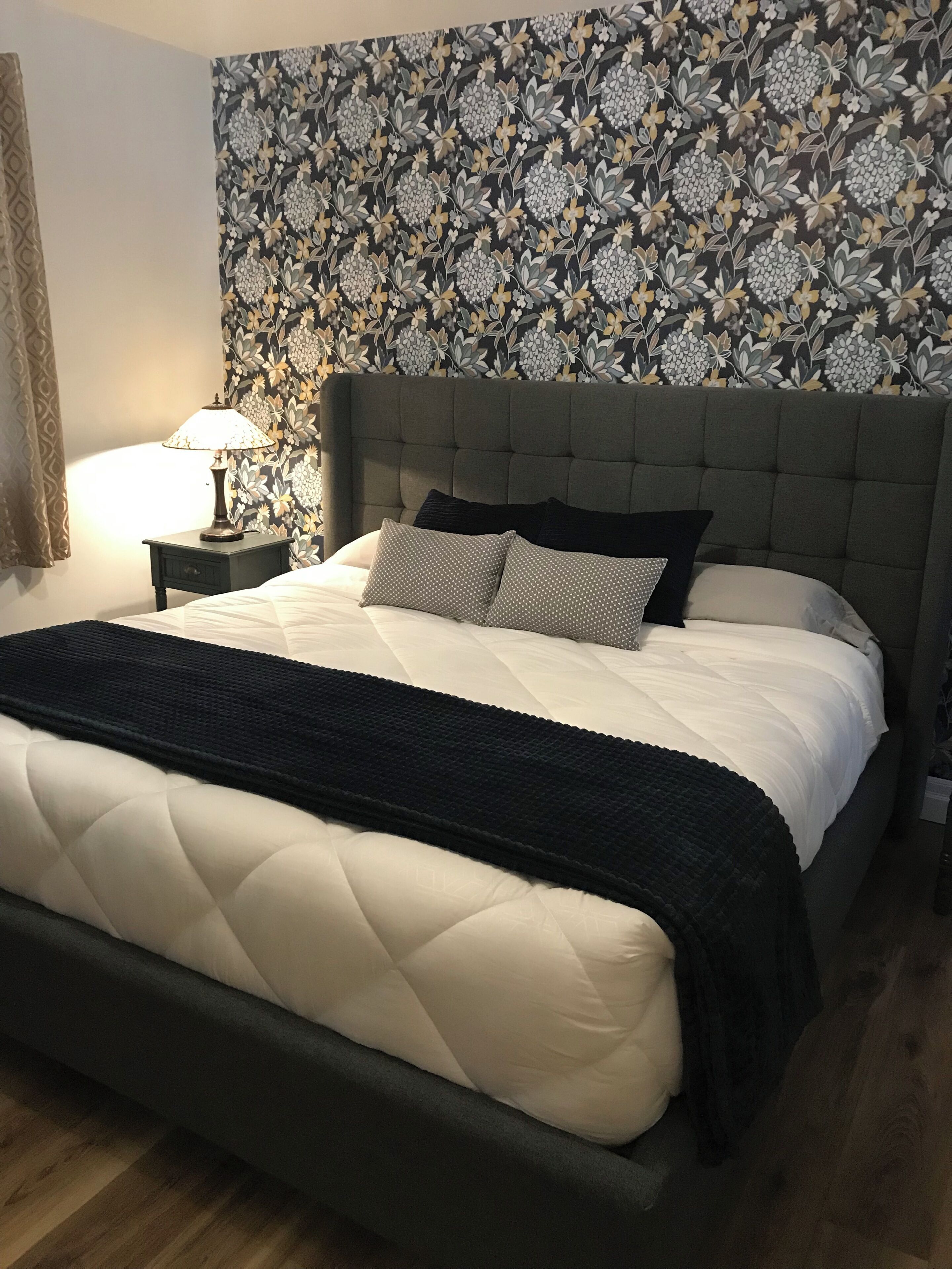 Una cama con cabecera capitonada, una mesita de noche con lámpara y papel tapiz estampado.