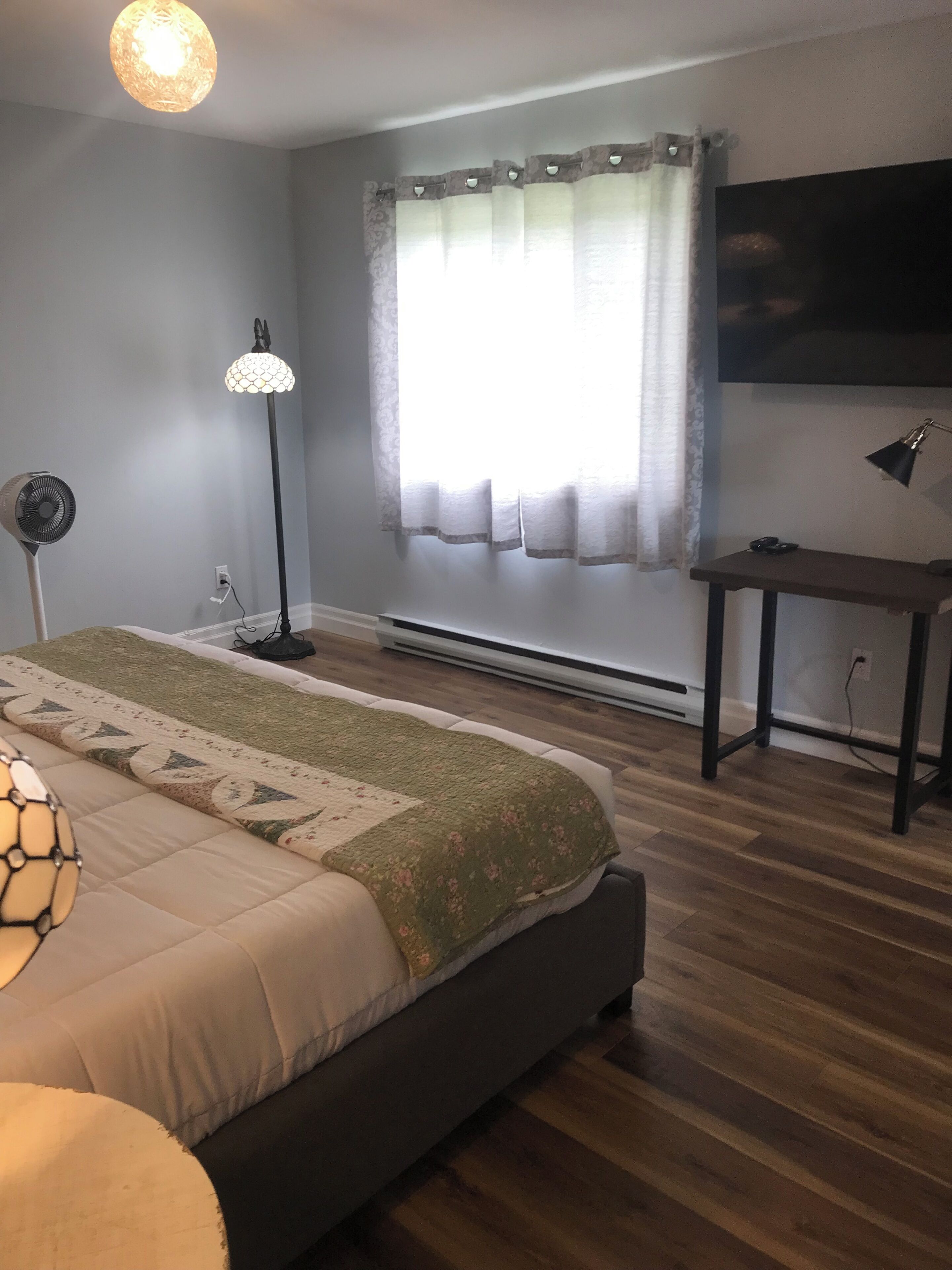 Un dormitorio con cama, lámpara de pie, lámpara de suelo, escritorio y ventana con cortinas.