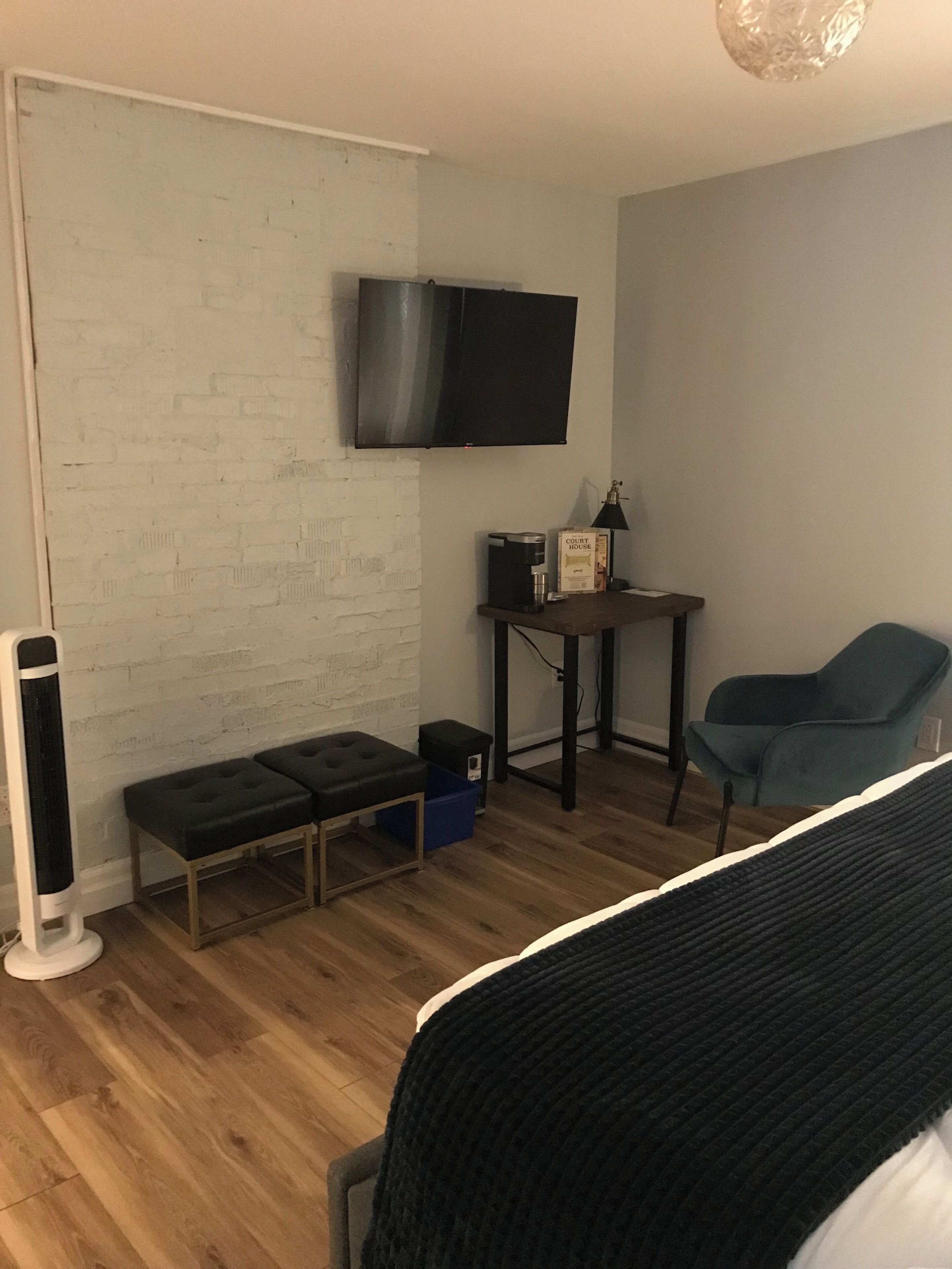 A room with a bed, a bench, a table with a coffee maker, a chair, and a TV mounted on the wall.